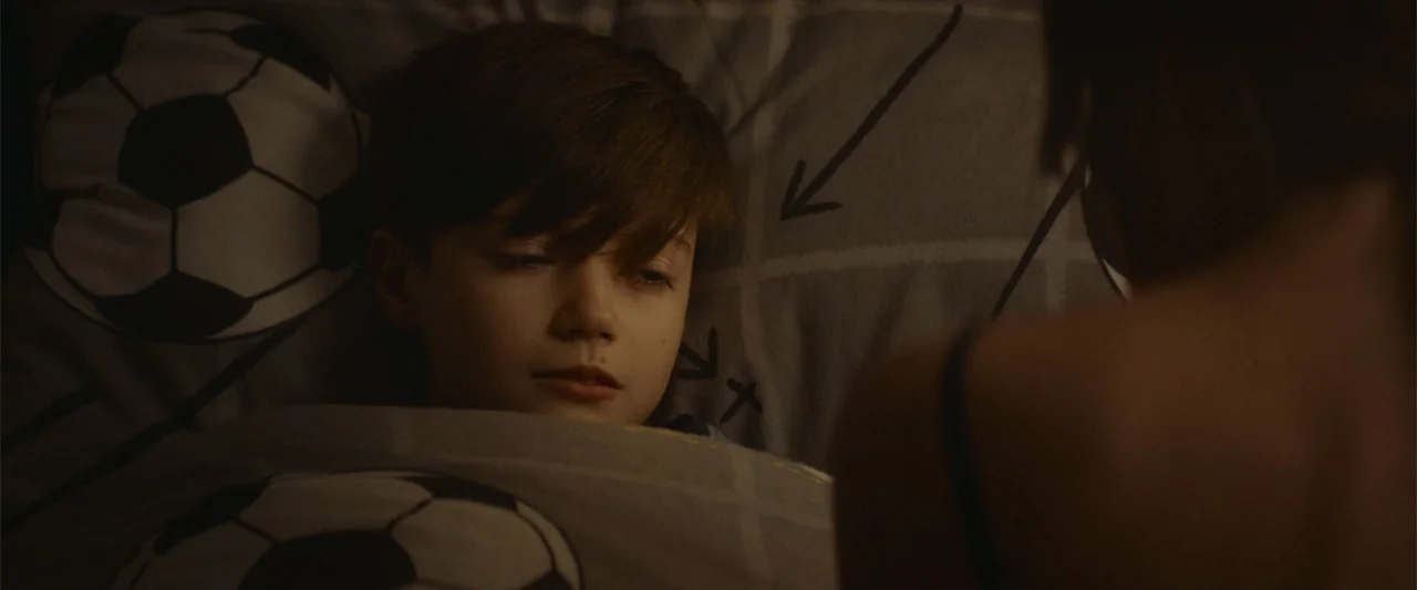 The Cellar (2022) movie still: closeup — A dimly lit bedroom features a child lying under patterned soccer-themed bedding. A blurred figure sits in…
