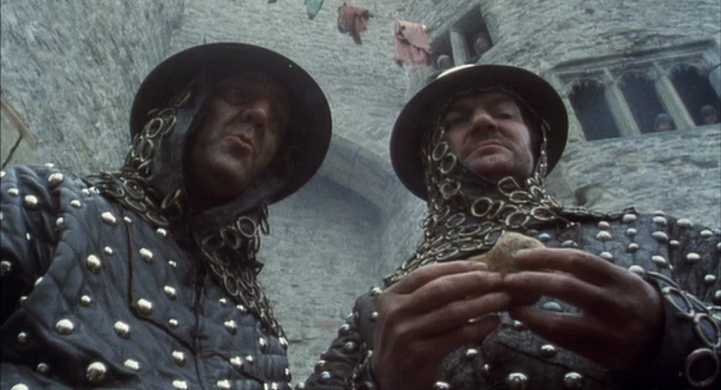 Jabberwocky (1977) movie still: medium — A stone courtyard with laundry hanging from a line above against a castle wall. Two men in medieval armor…