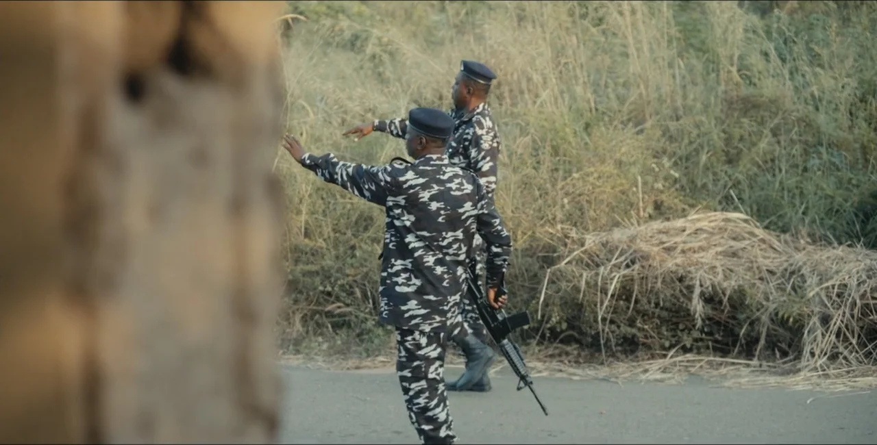 Project Assault (2025) movie still: fullbody — Two uniformed officers stand on a roadside bordered by dry tall grass. They gesture toward the…