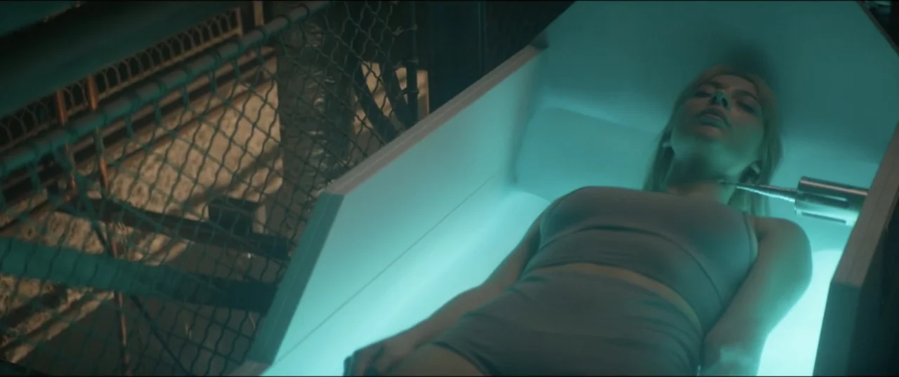 Utopia (2013) movie still: medium — A sleek white medical stasis pod is situated in a dark industrial space with wire fencing. A young woman lies…