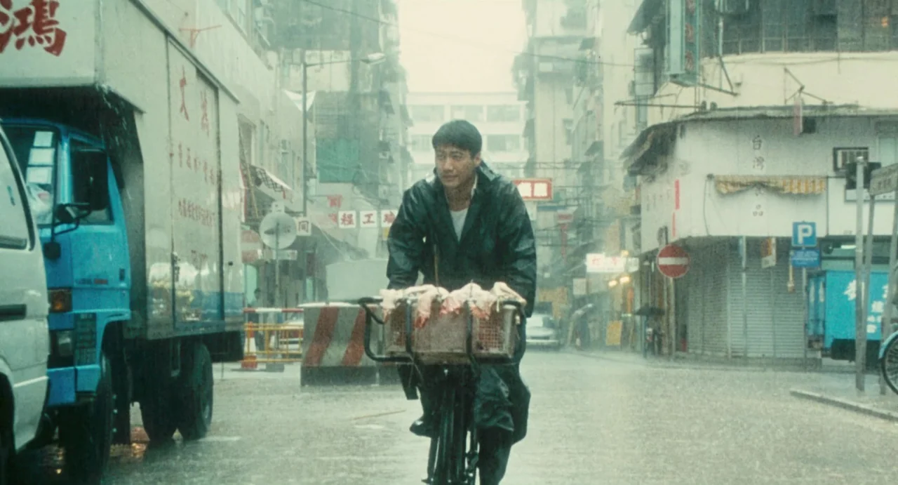 Comrades: Almost A Love Story (1996) movie still: fullbody — A wet urban street in Hong Kong is filled with rain and older vehicles. A man rides a…