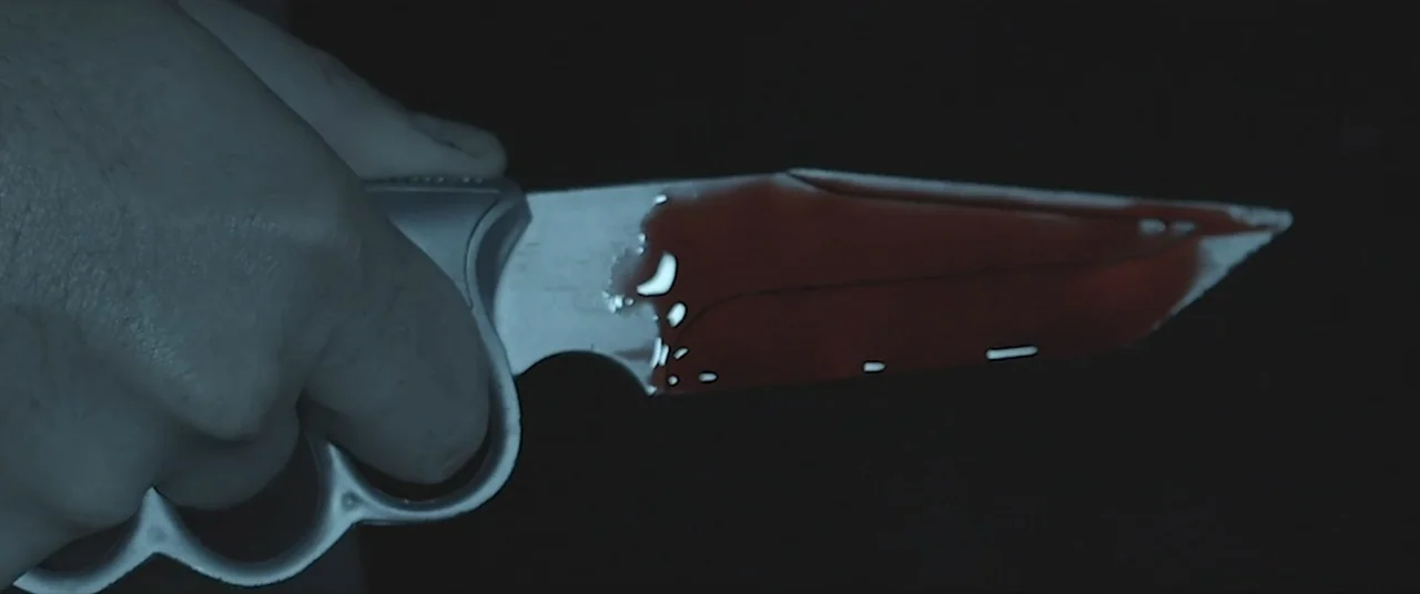 Amityville Cult (2021) movie still: closeup — A dark, nondescript interior space features a close view of a weapon. A hand grips a steel trench…