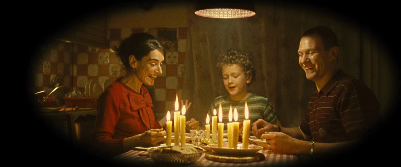 Micmacs (2009) movie still: medium — A dimly lit room features a family dining at a table illuminated by candles. The group laughs and eats together…