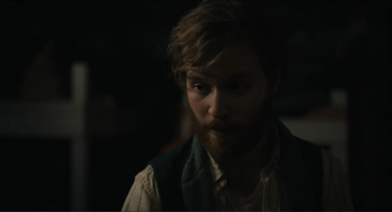 Hell of a Summer (2025) movie still: closeup — A dark, dimly lit room contains a man. The man with short wavy hair sits facing three-quarter.