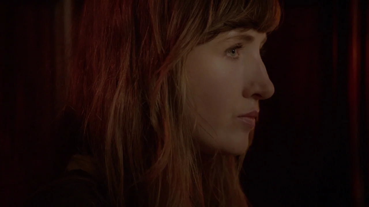 The Heart Machine (2014) movie still: closeup — A dark, dimly lit indoor space with wood-paneled walls. A woman with shoulder-length brown hair…