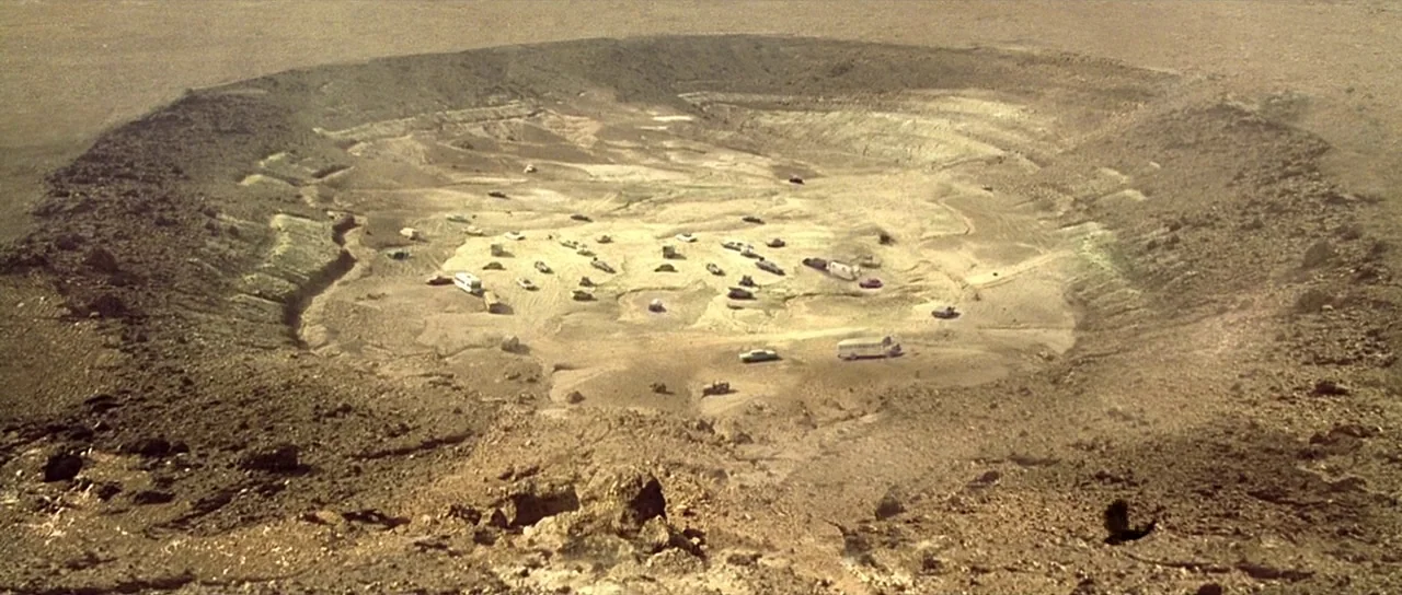 The Hills Have Eyes (2006) movie still: establishing — A vast, barren desert crater holds a scatter of various automobiles and trailers inside. The…
