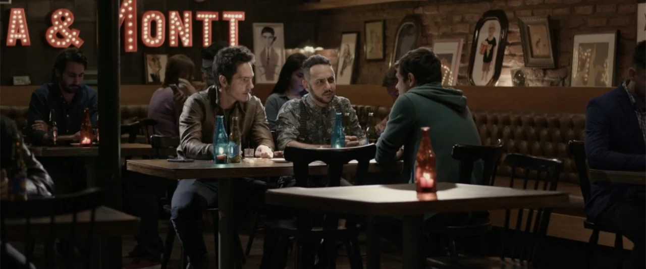 Do It Like An Hombre (2017) movie still: medium — A dimly lit bar with brick walls and framed artwork decorates the background. Three men sit at a…