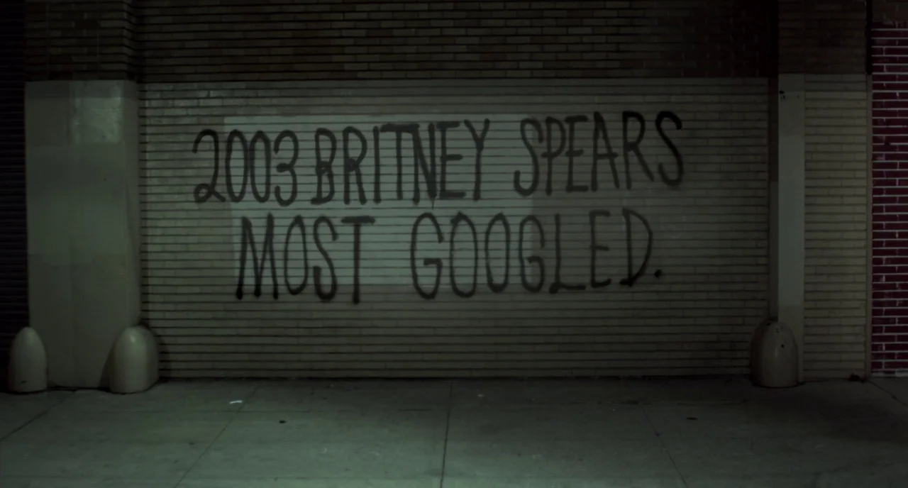 Beginners (2010) movie still: establishing — A dark brick wall at night features spray-painted black lettering on a lighter patch. The text reads…