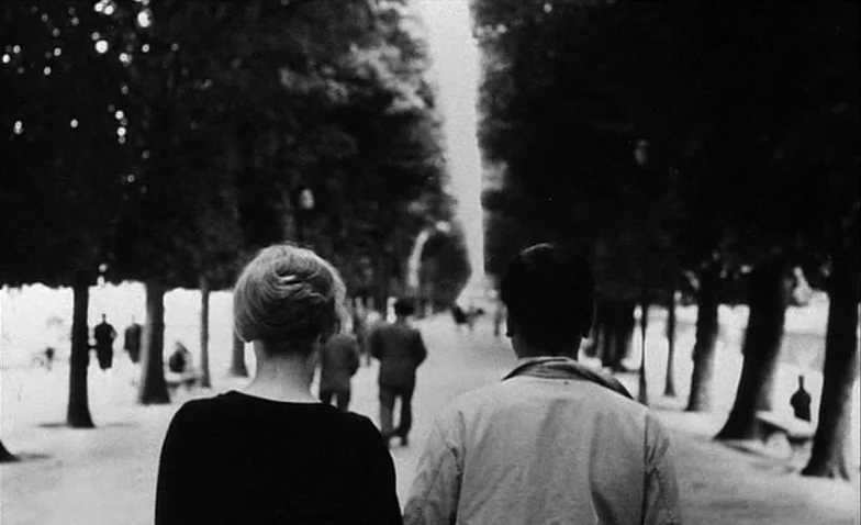La Jetée (1962) movie still: medium — A tree-lined walkway in a park with multiple distant figures visible along the path. Two people walk away from…