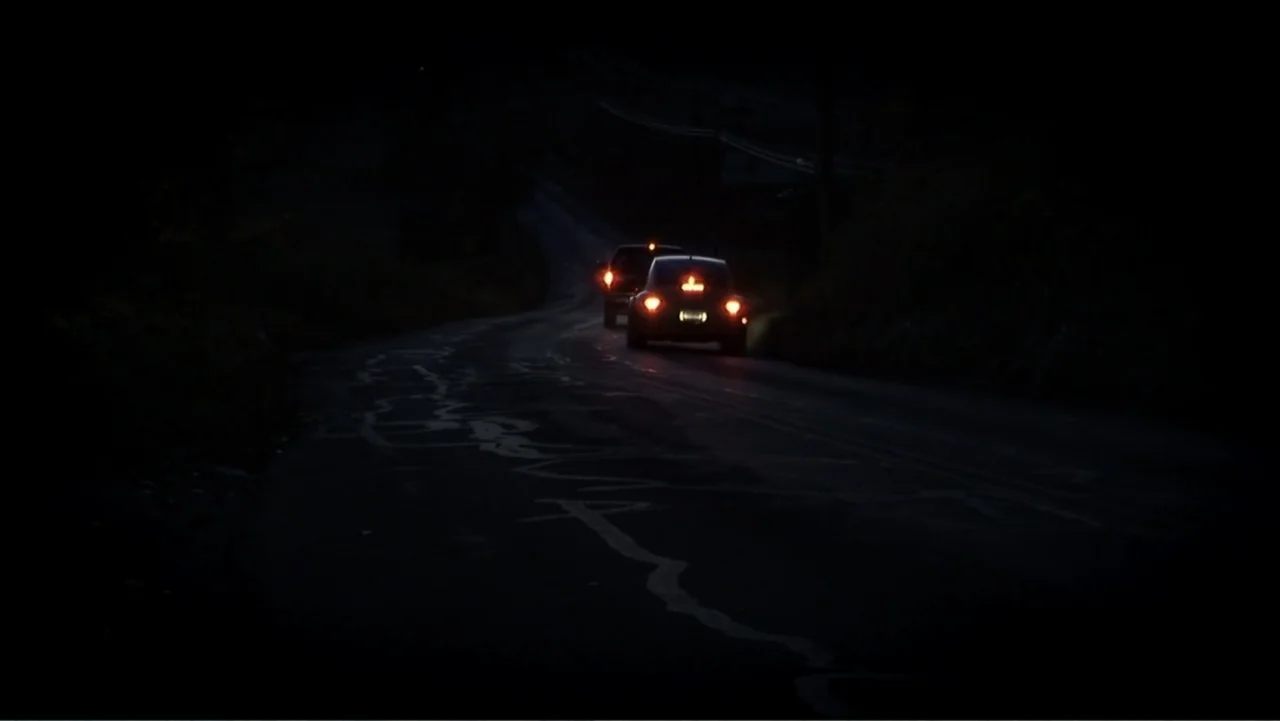 The Cheshire Murders (2013) movie still: wide — A dark, winding road extends through a shadowed landscape at night. Two cars drive away from the…