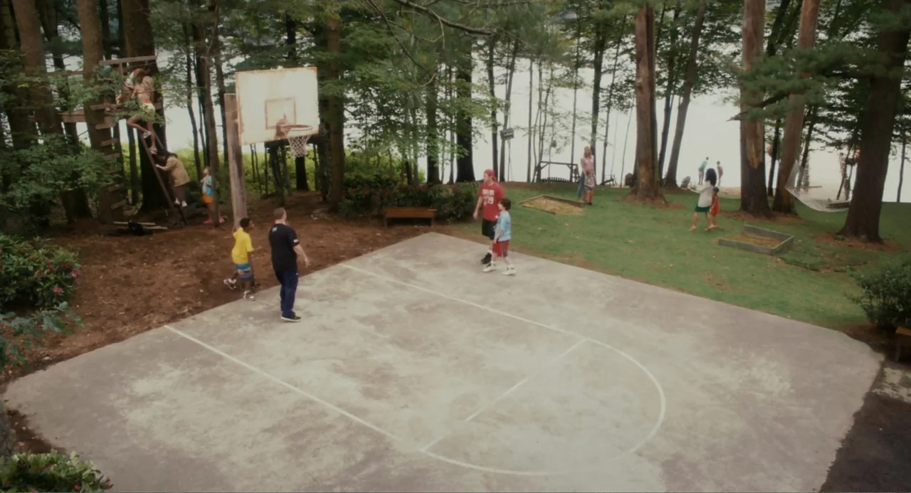 Grown Ups (2010) movie still: wide — A concrete basketball court is situated in a wooded area beside a calm lake. Several people are engaged in…