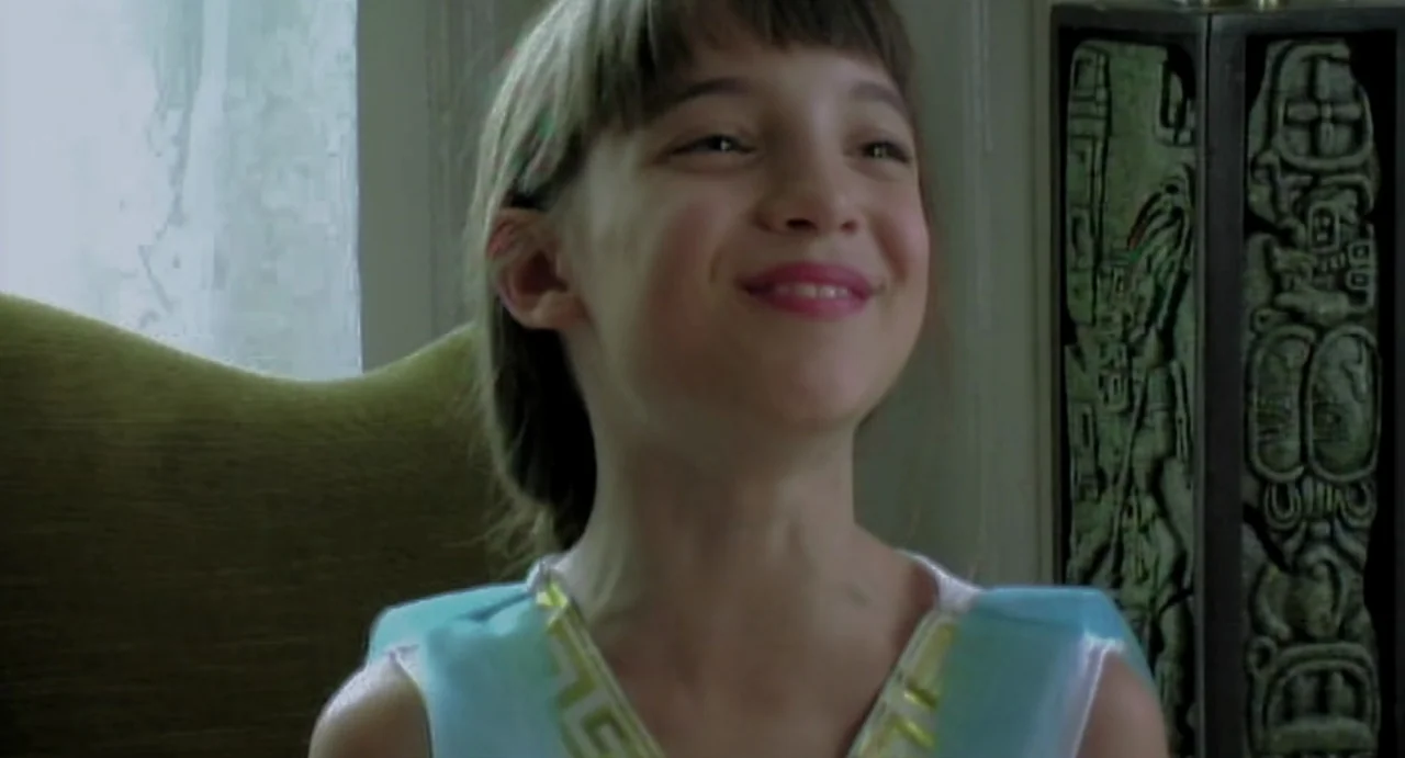 La Abuela (2021) movie still: closeup — An interior space features a green chair and a textured green decorative column. A young girl in a blue…