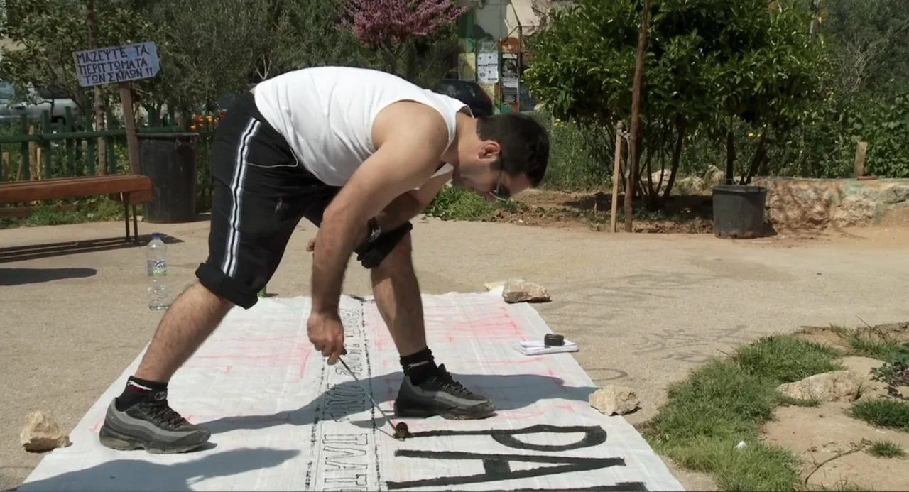 Az X- Kryptonit projekt (2020) movie still: fullbody — A man bends over a long white banner laid flat on the ground. He is using a brush to paint…