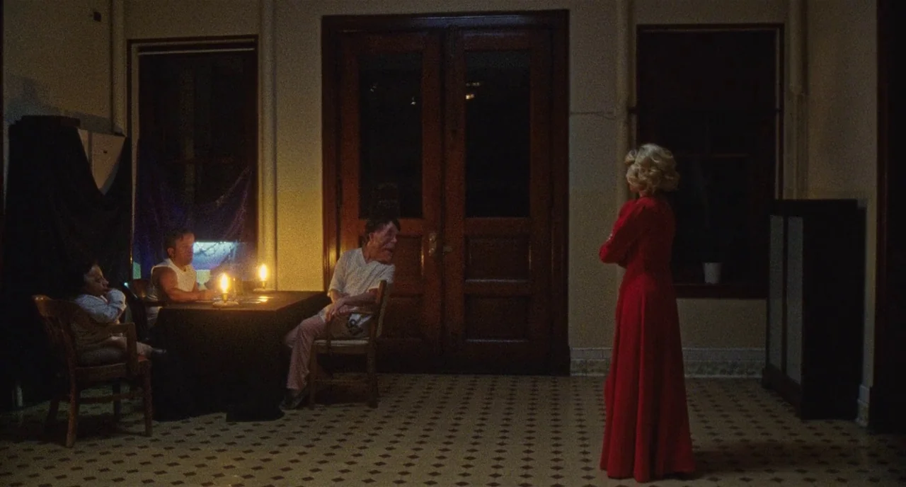 Chained For Life (2018) movie still: wide — A dimly lit room features a checkerboard tile floor and large wooden double doors. A woman in a red gown…