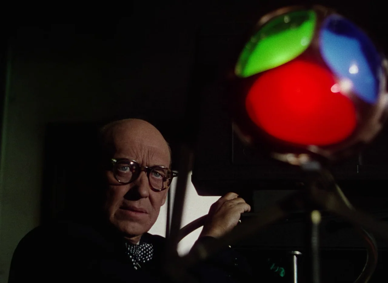 The War of the Worlds (1953) movie still: closeup — A dimly lit indoor space with a man positioned near technical filming equipment. The man gazes…