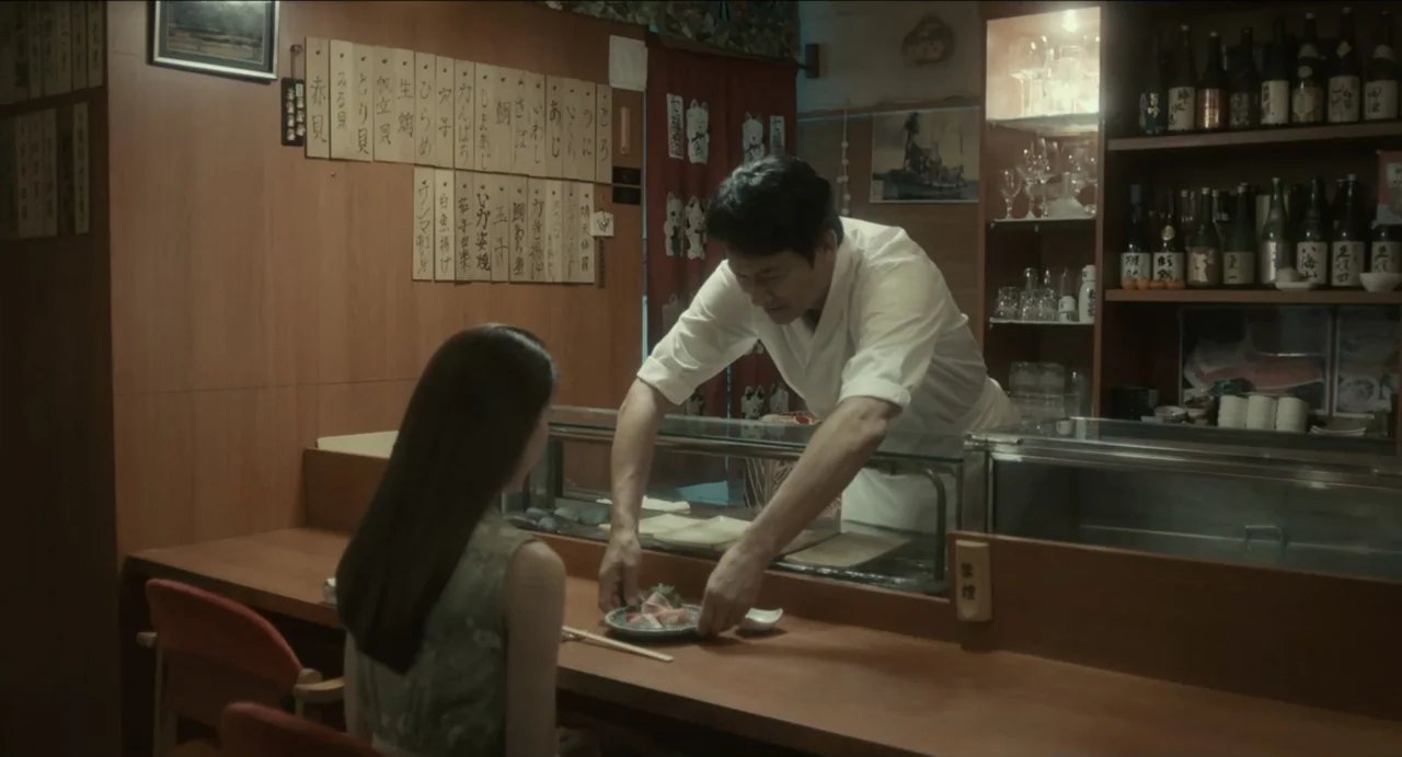 Ramen Shop (2018) movie still: medium — A small sushi restaurant interior with wooden walls featuring hanging Japanese menu placards. A chef in a…