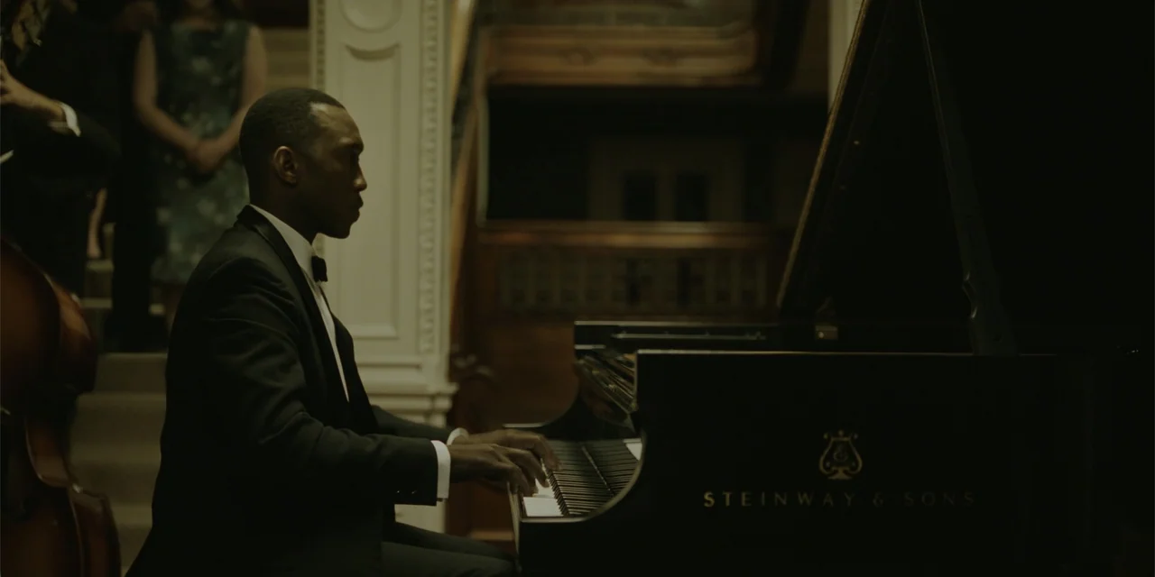 Green Book (2018) movie still: medium — A dark indoor space features a grand piano and a wooden staircase. A man in a formal tuxedo sits at the…