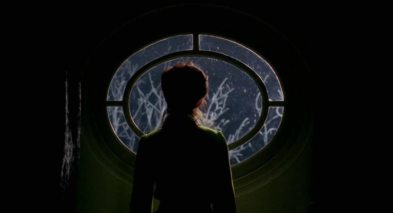 8 Women (2002) movie still: medium — A dark hallway contains a circular window looking out onto bare winter tree branches. A woman stands in front…