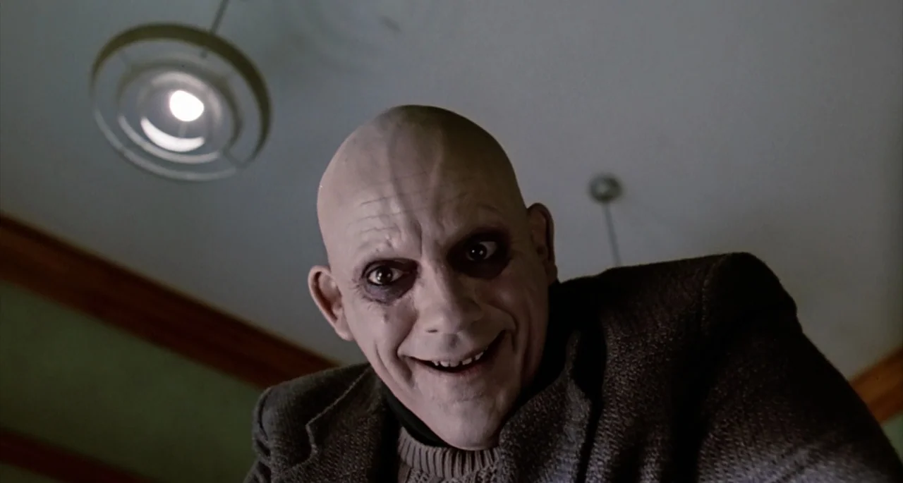 The Addams Family (1991) movie still: closeup — A dimly lit indoor space featuring a simple overhead ceiling light fixture. A bald man with pale…