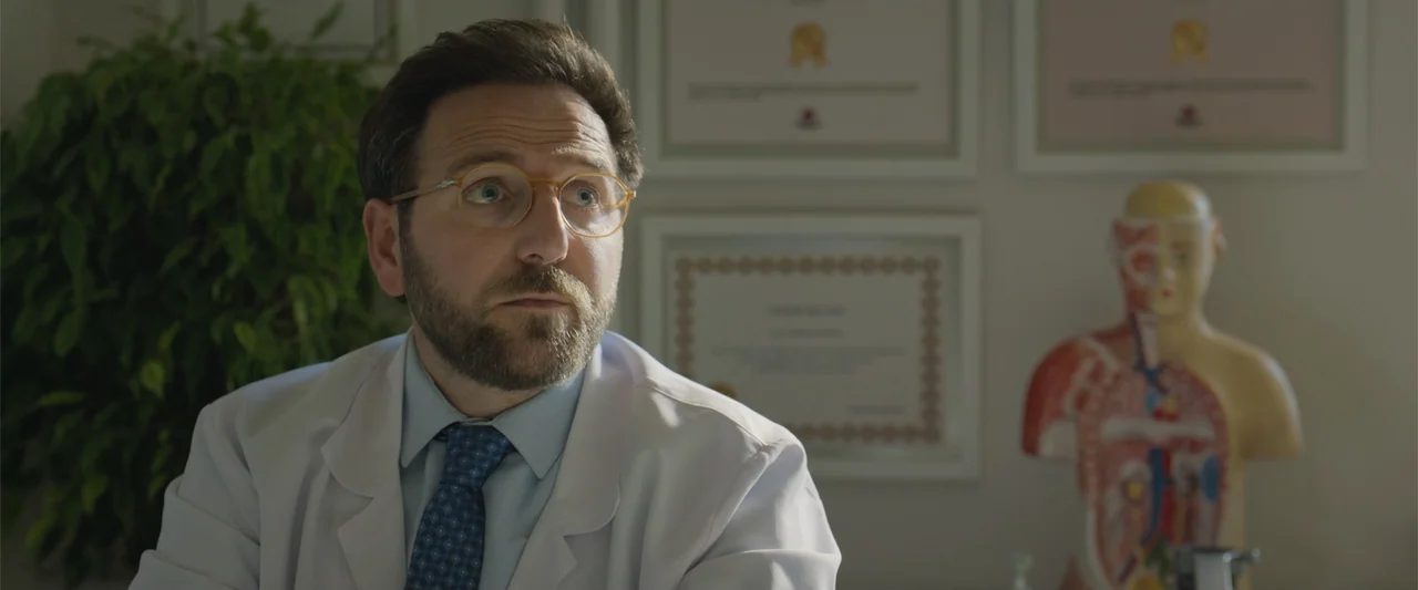 Blue Cave (2024) movie still: medium — A medical office with framed degrees on the wall and an anatomical torso model. A bearded male doctor in a…