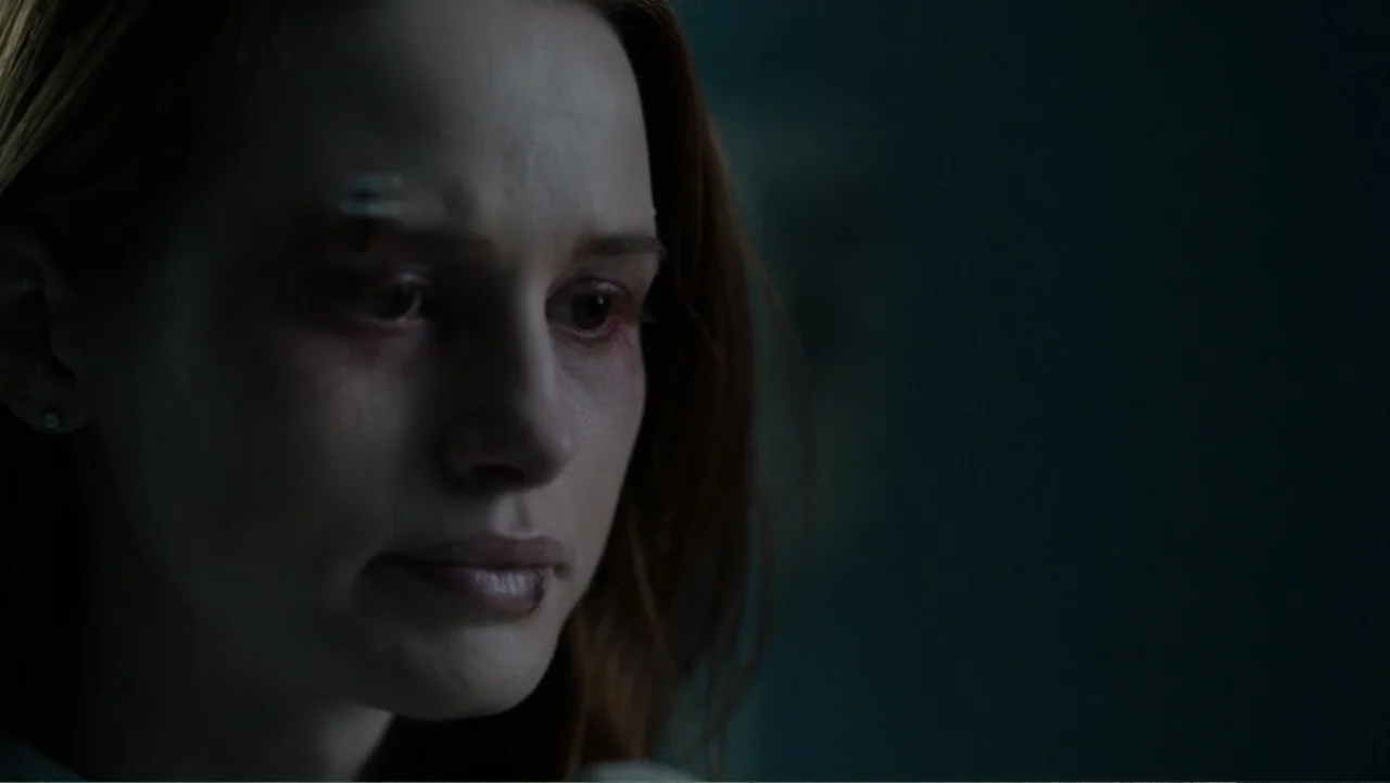 Sightless (2017) movie still: closeup — A dimly lit room with muted blue tones and indistinct architectural features in darkness. A young woman with…