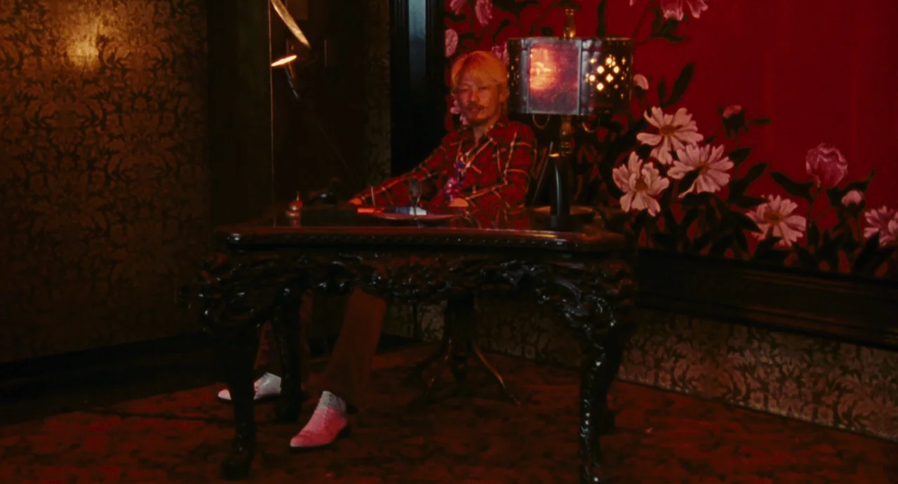 Ichi The Killer (2001) movie still: medium — A dark, dimly lit study features ornate floral patterned red wallpaper and dark furniture. A blonde man…