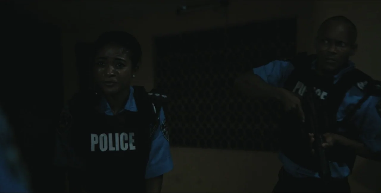 Project Assault (2025) movie still: medium — A dark, sparsely furnished interior with a textured wall pattern visible in the shadows. Two police…