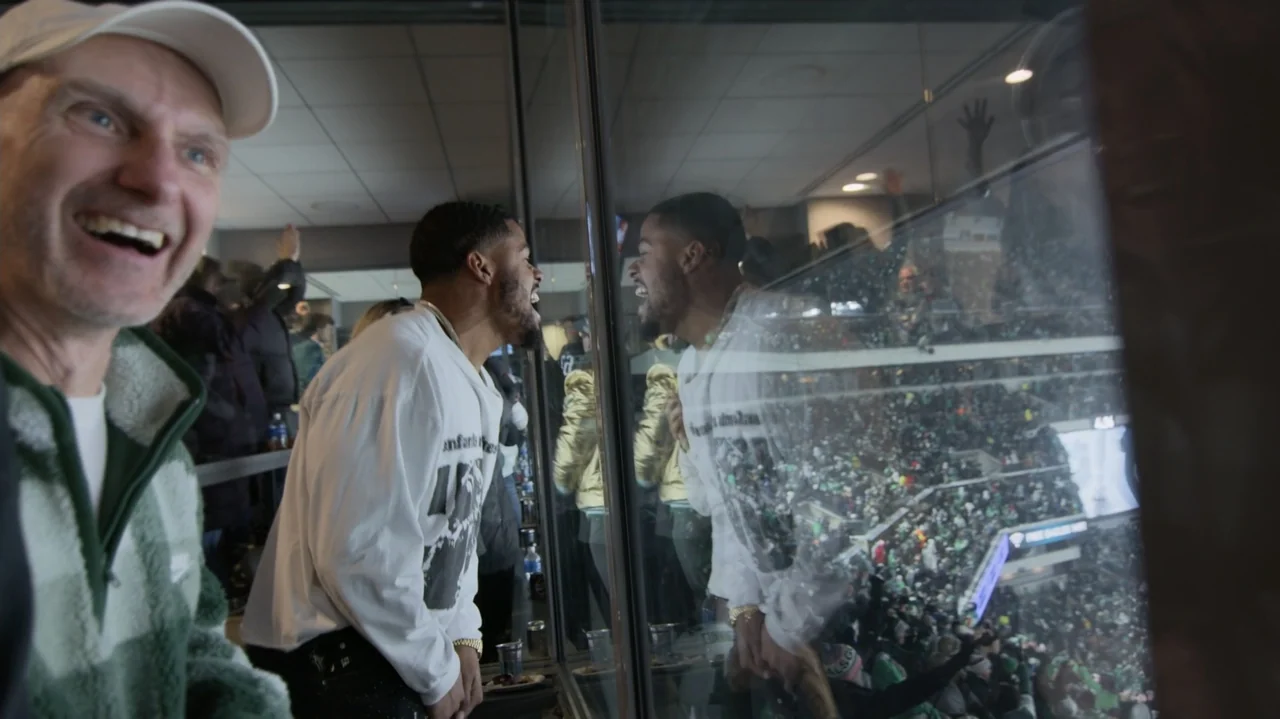 SAQUON (2025) movie still: medium — An interior luxury suite with glass windows overlooks a large crowded stadium at night. Two men stand near the…