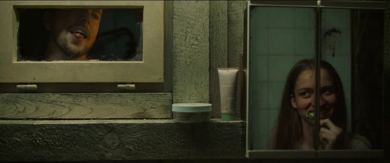 Nightsiren (2022) movie still: medium — A dimly lit room with textured wooden walls and a small rectangular window. A woman brushes her teeth in a…