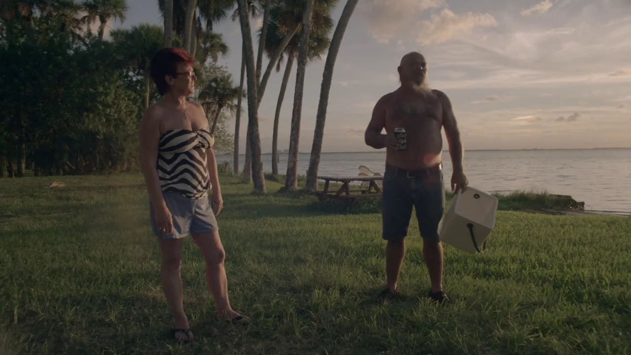 Florida Man (2015) movie still: fullbody — A grassy area populated by palm trees sits adjacent to a calm lake. A man and a woman stand together on…