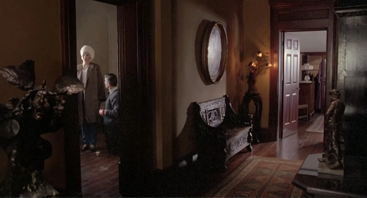 Dead of Winter (2025) movie still: medium — A dimly lit residential corridor features hardwood floors and ornate antique furniture pieces. A man and…