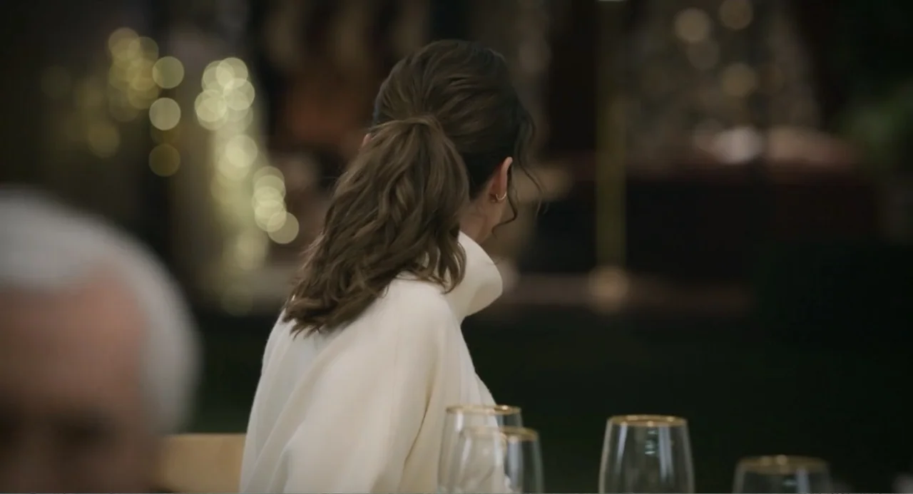 La posada (2023) movie still: medium — An interior dining area with blurred festive lights visible in the out-of-focus background. A woman in a…