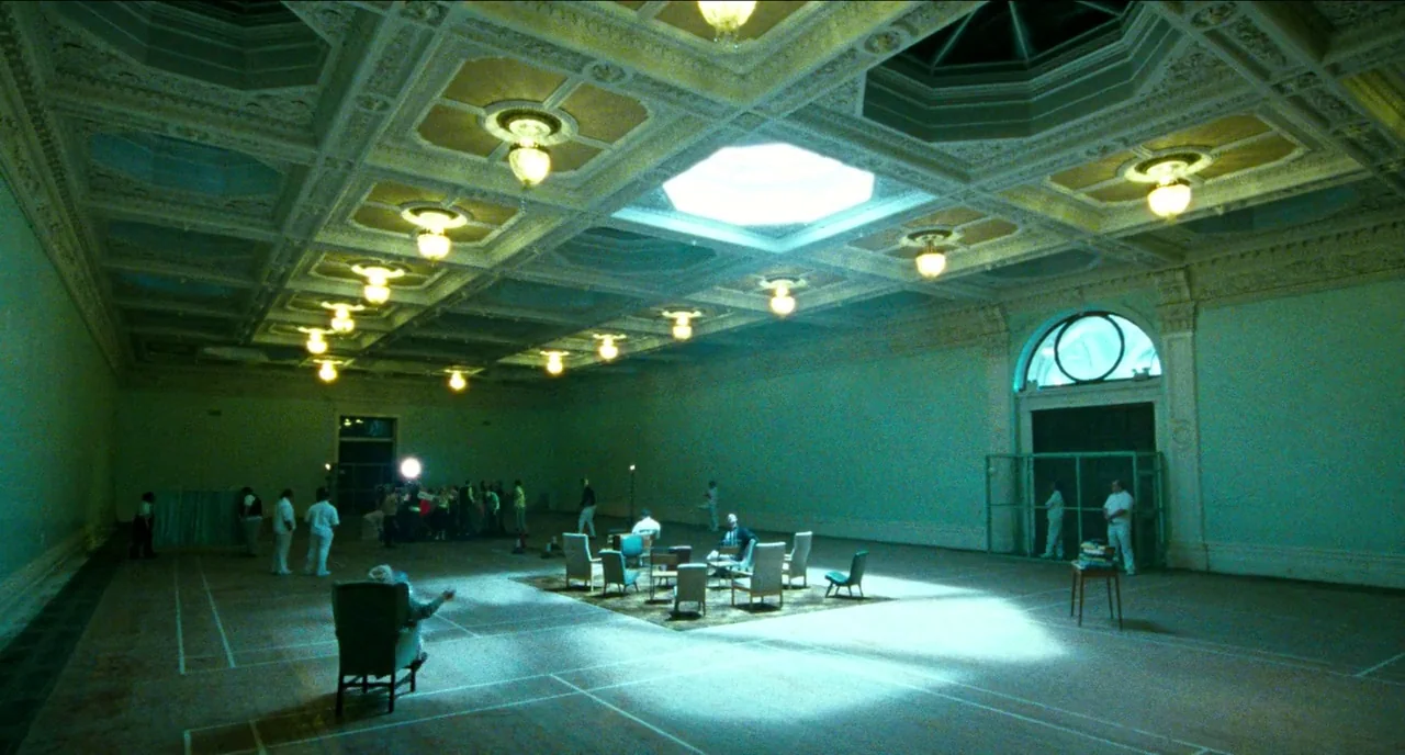 Bronson (2008) movie still: wide — A vast, high-ceilinged room features intricate white molding and a central light-filled skylight. Several people…
