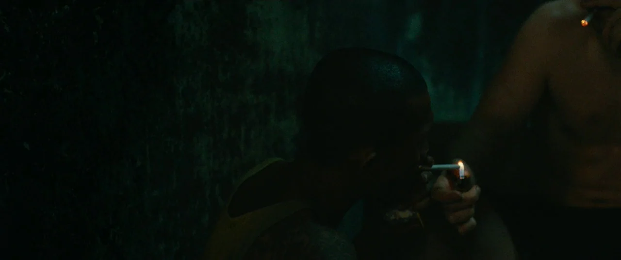 A Prayer Before Dawn (2017) movie still: medium — A dark, cramped interior with textured walls and low visibility. Two shirtless men light…