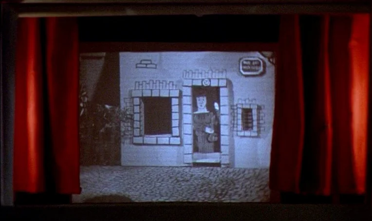 Jacquot de Nantes (1991) movie still: wide — A small two-dimensional puppet house sits on a stage behind bright red velvet curtains. A simple figure…