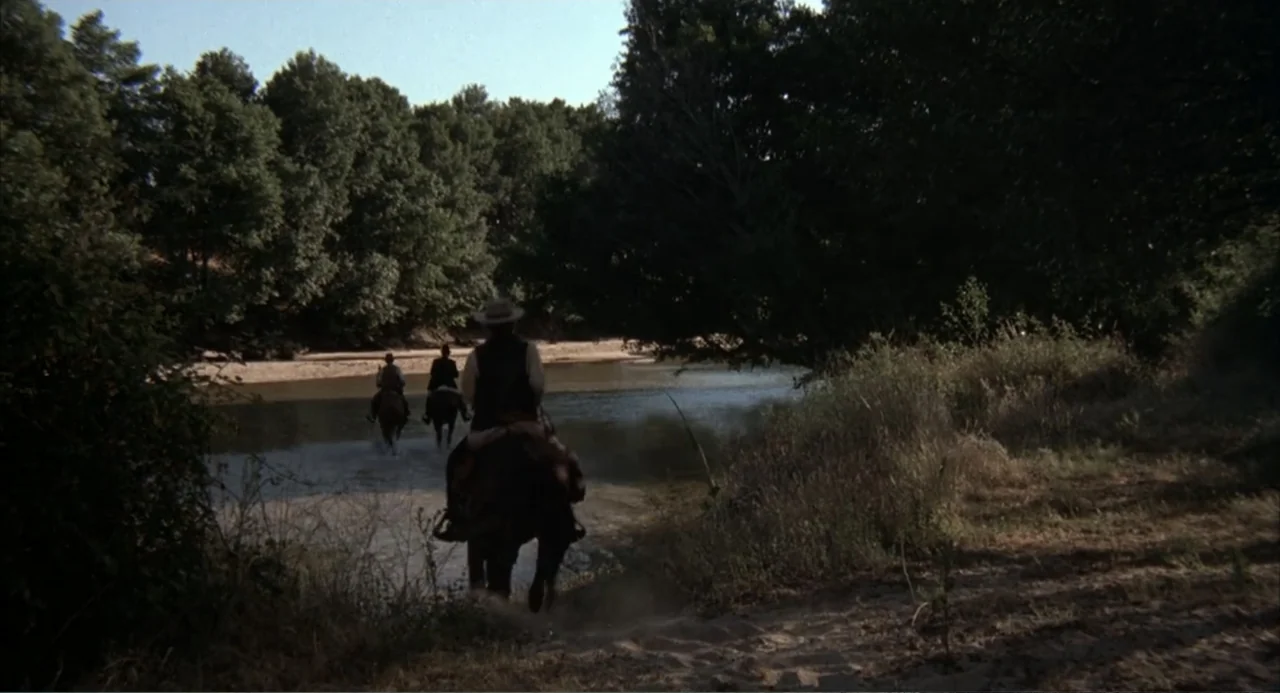 The Spikes Gang (1974) movie still: wide — Three riders on horseback cross a shallow river bordered by dense green trees. The scene is shot from a…