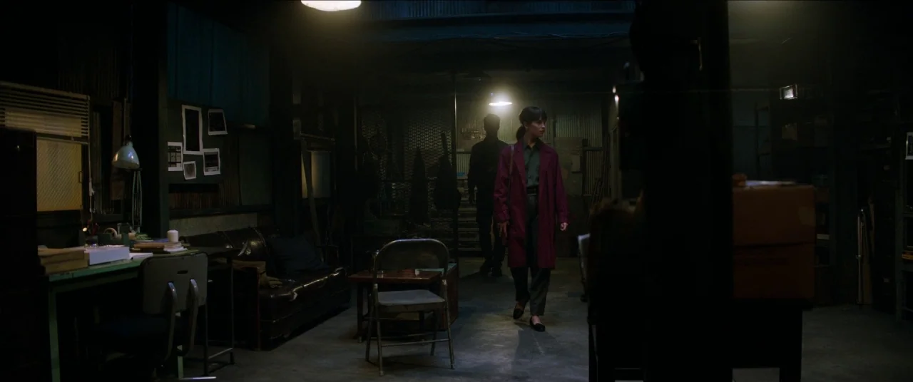 Earthquake Bird movie still: wide — A dimly lit room functions as an austere office space with metal walls. A woman in a burgundy coat walks toward…