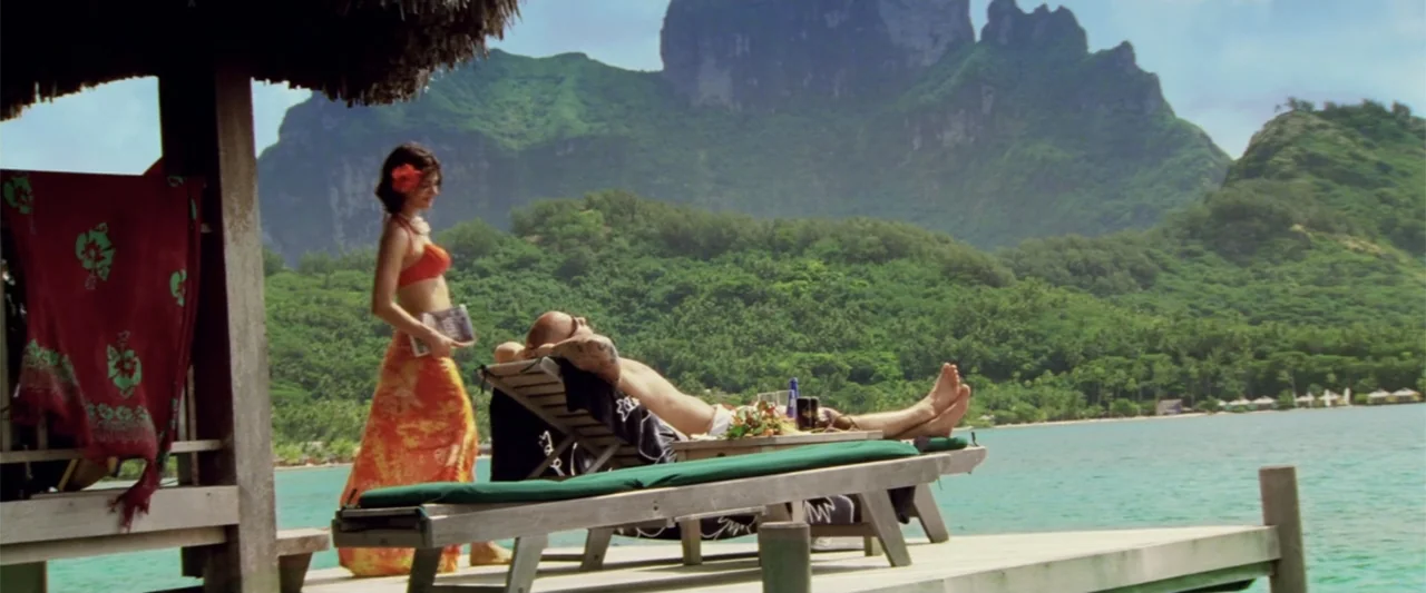 xXx (2002) movie still: fullbody — A wooden dock extends over turquoise water with a tropical island in the background. A man relaxes on a lounge…
