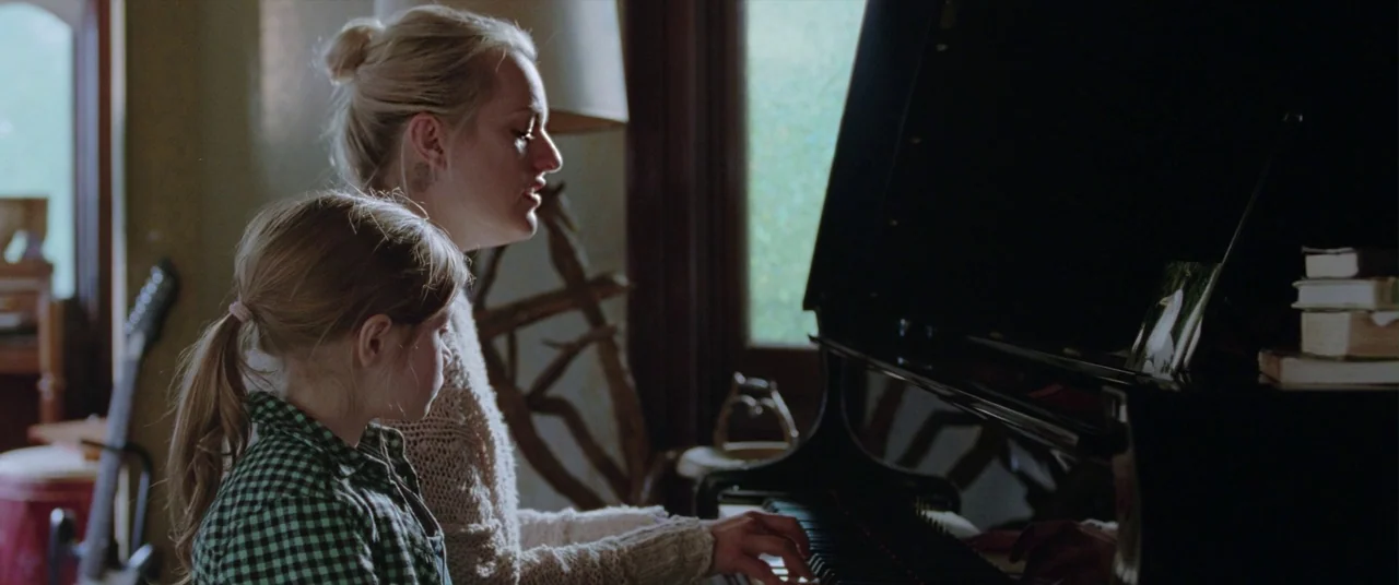 Her Smell (2018) movie still: medium — A dimly lit room features a black upright piano against a windowed wall. An adult woman and a young girl sit…