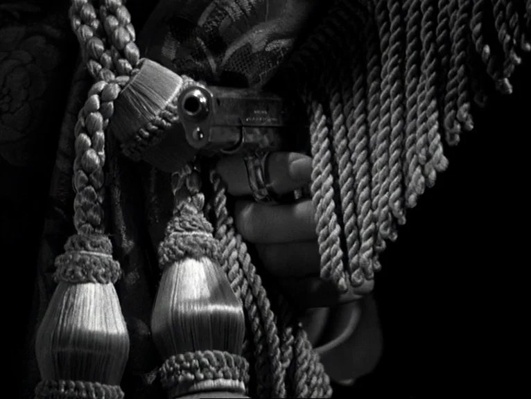 Saboteur (1942) movie still: closeup — A dark, textured fabric curtain with thick braided fringe hangs in a black space. A hand holds a small…