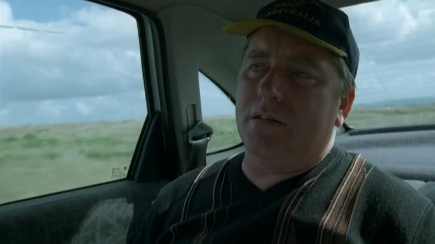 Garage (2007) movie still: medium — The interior of a moving car shows a man sitting in the passenger seat. Through the window, a flat, grassy…