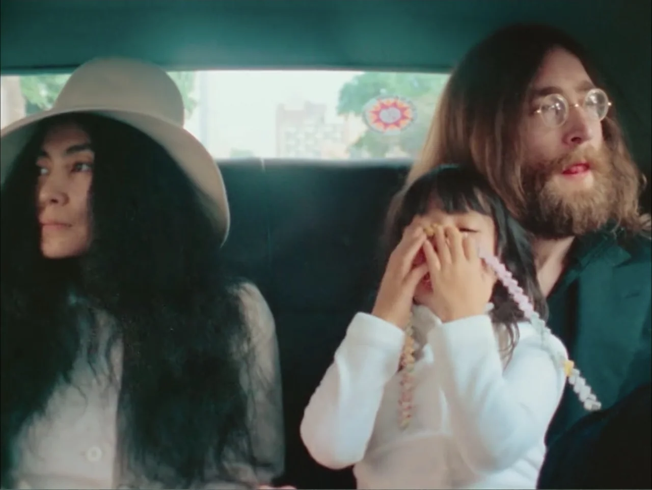 One to One: John & Yoko (2025) movie still: medium — Three people sit together in the backseat of a moving car during the day. A woman, a man with a…