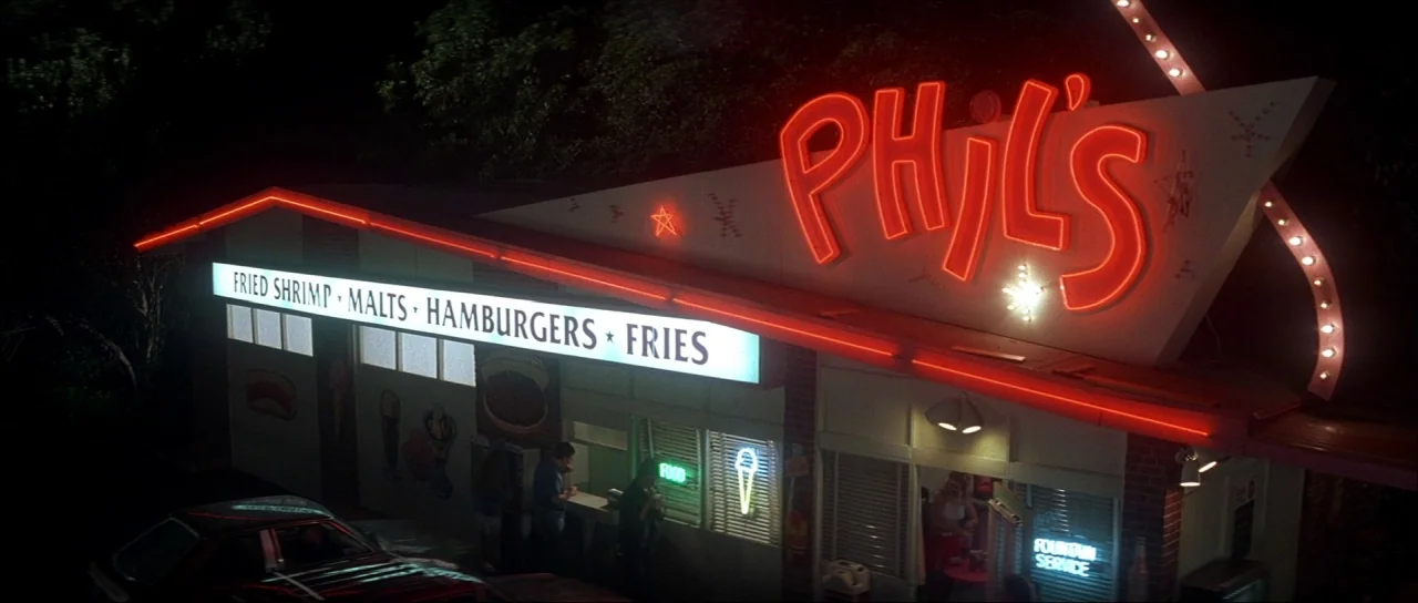 The Monster Squad (1987) movie still: establishing — A roadside diner at night features a prominent red neon sign and illuminated menu. The…