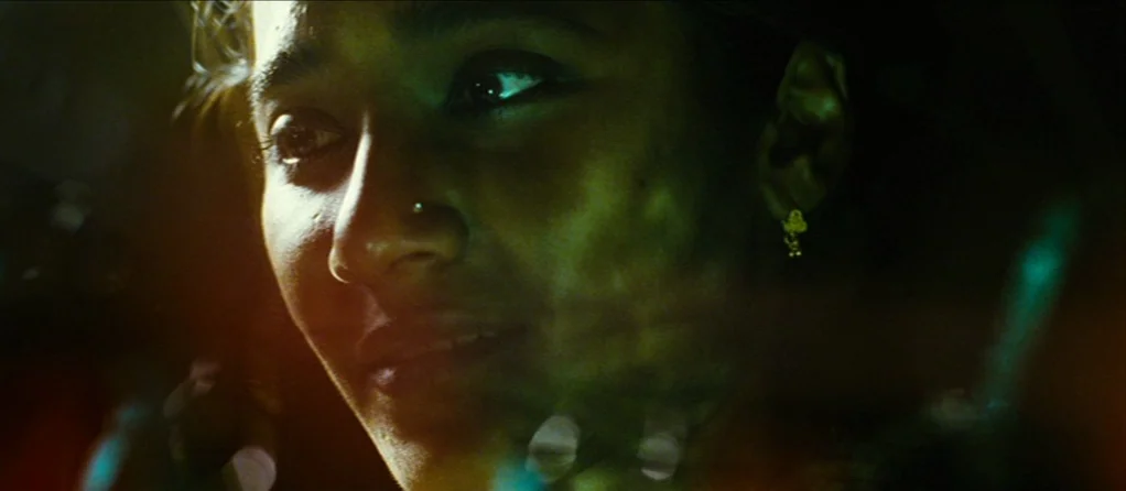 Brick Lane (2007) movie still: closeup — A dark vehicle interior at night is illuminated by flickering neon external lights. A young woman with a…