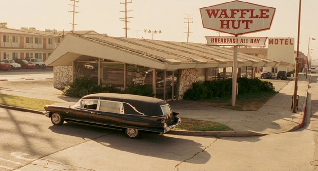 The Ladykillers (2004) movie still: wide — A low-slung Waffle Hut building sits beside a motel under clear skies. A vintage black hearse is parked…