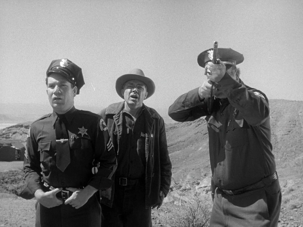 The Prowler (1951) movie still: medium — A dry, rocky desert landscape stretches out under a clear, bright daylight sky. Two uniformed police…