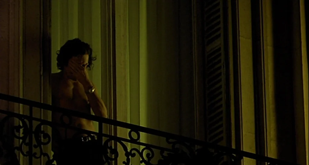 Trouble Every Day (2001) movie still: medium — A dark balcony at night features wrought iron railings and a tall glass doorway. A shirtless man…