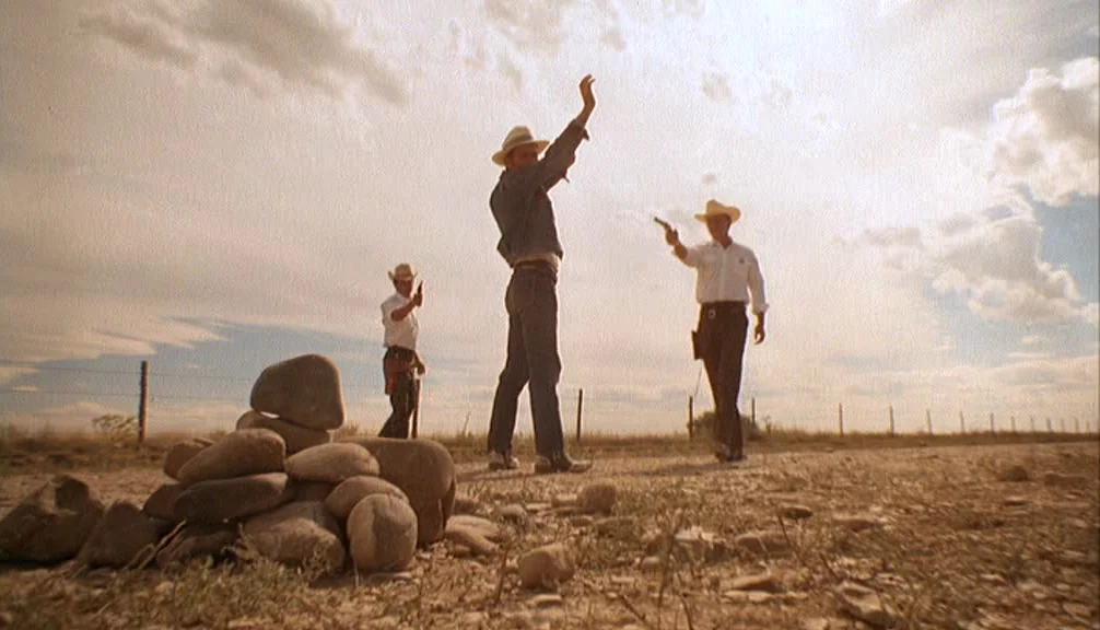 Badlands (1973) movie still: wide — An arid, flat desert landscape features a dirt road bisected by a wire fence. Three men in cowboy hats hold…