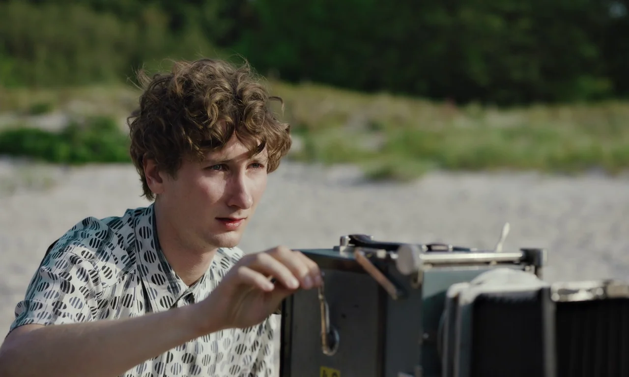 Bloodsuckers (2021) movie still: medium — A man sits on a sandy beach behind a large antique bellows camera. He carefully turns the metal focus dial…