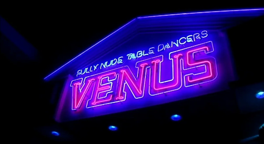9 Songs (2004) movie still: establishing — A dark exterior facade is dominated by a large glowing neon sign for a venue. The sign displays the name…